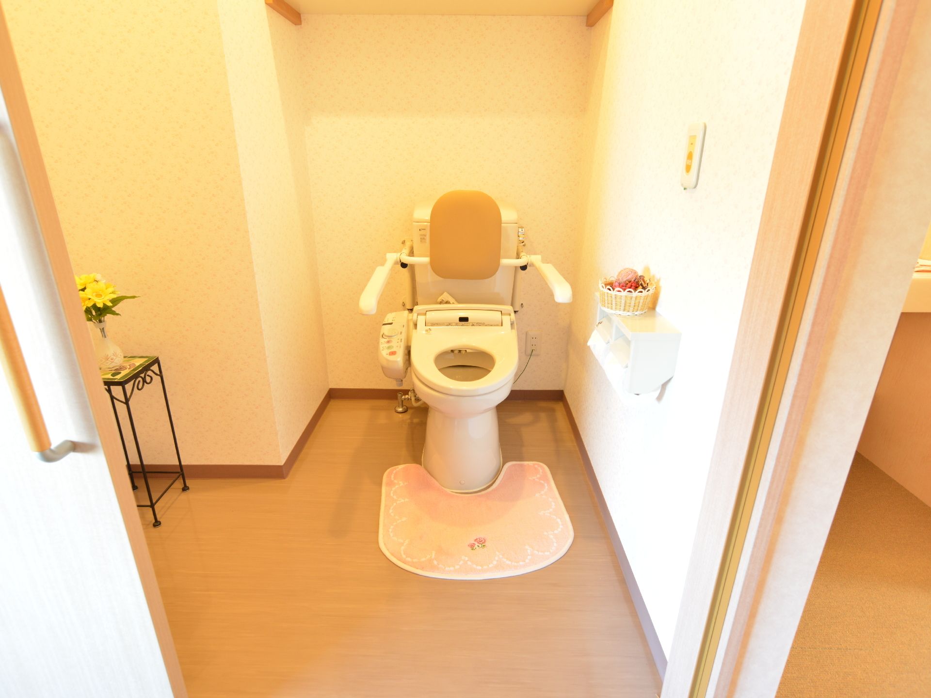 広めのトイレはウォッシュレットがついている。お花が飾られている。トイレは手すりと背もたれがついている。