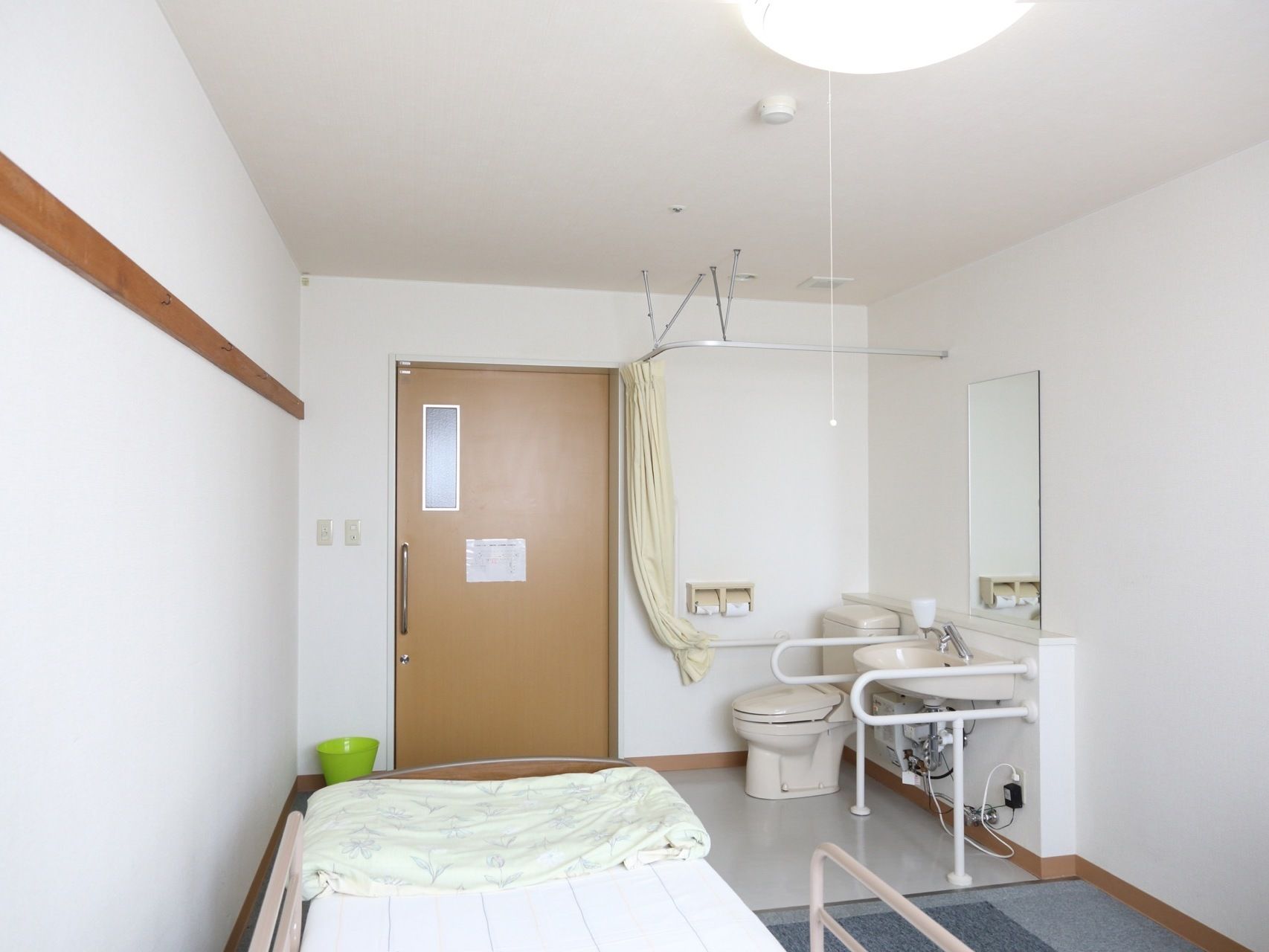 居室の手前には、幅が広いベッドがあり、壁際にはドアがなく、オープンスペースとなっている便座と手すりが設置されている。