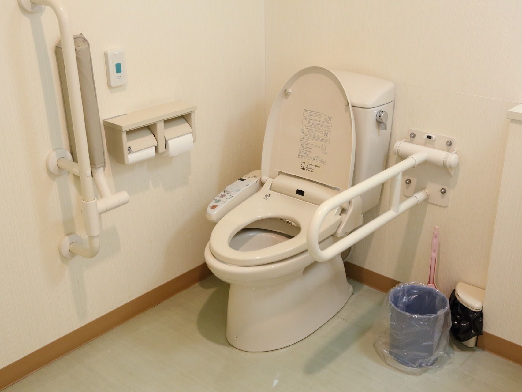 手すり付きの便座の横には、トイレットペーパー２つと、ナースコールが壁際に設置されている。床部分にはゴミ箱もある。
