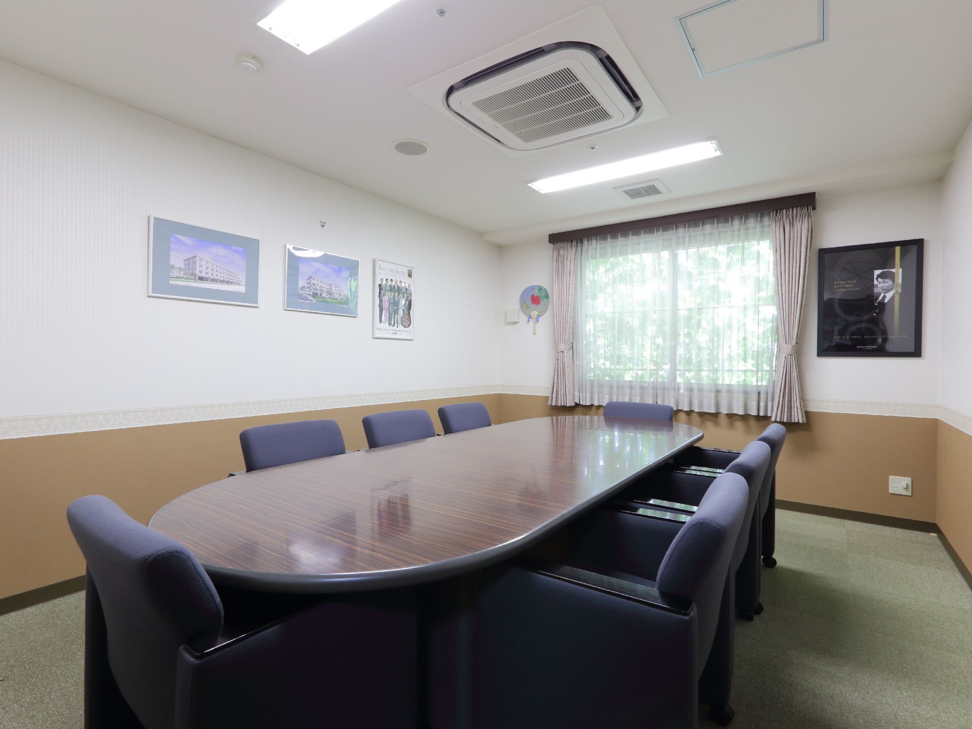 応接室には天井にエアコンが設置されており、丸い長テーブルと椅子が8脚置かれています。壁には、額に入った写真が飾られている。