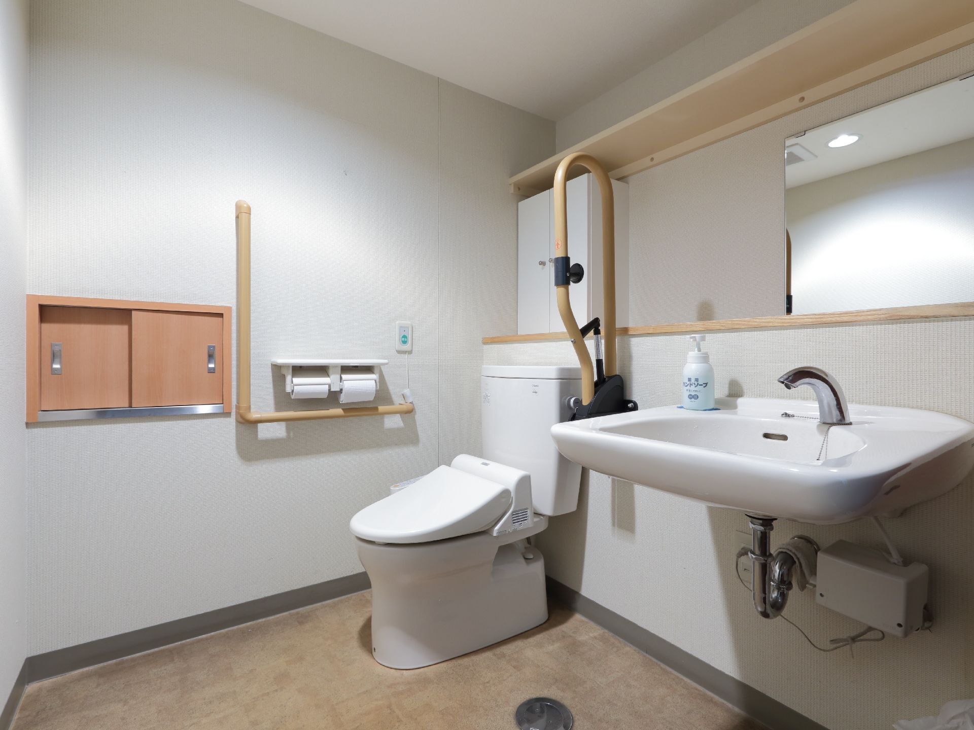 白い壁と天井、焦げ茶色の模様入り床のトイレ。便器の隣に洗面台がある。洗面台の上には鏡があり、便器の傍には手すりが設置。
