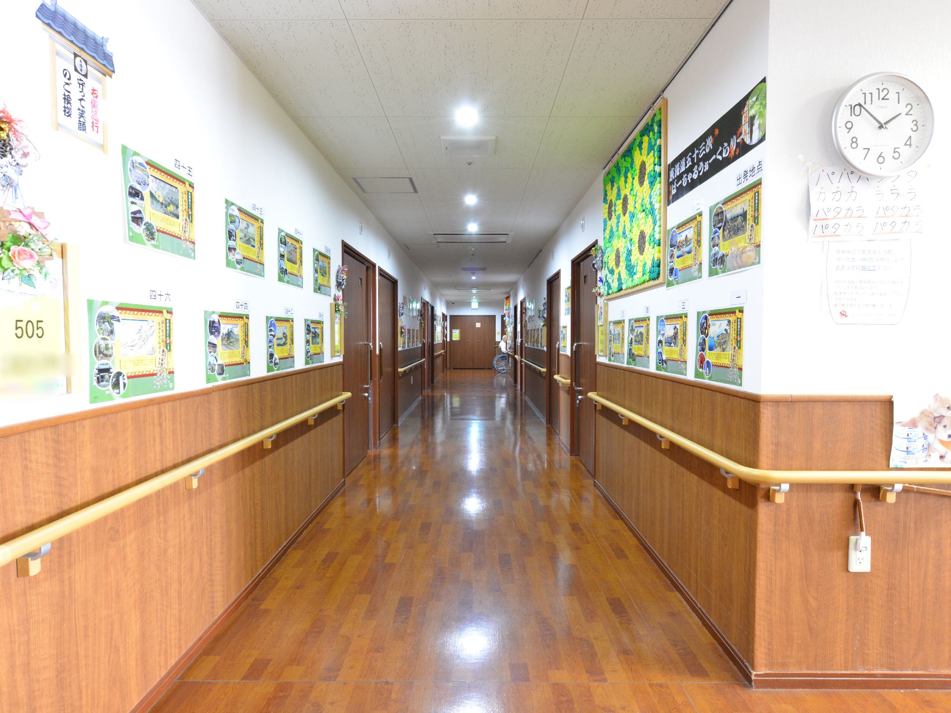 白と焦げ茶の木目調でまとめられた個室の通路。両側の壁には手すりが設置され、部屋のドア横には部屋番号が掲げられている。