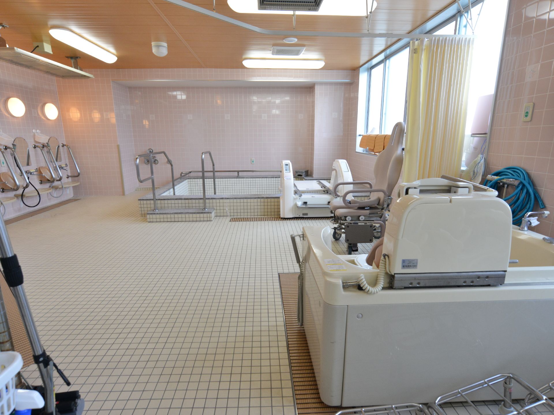 手前右側に入浴機器、その先と左側に洗い場、右奥に浴槽がある。右側が透明なガラス窓で光がよく入り、浴室全体が非常に明るい。