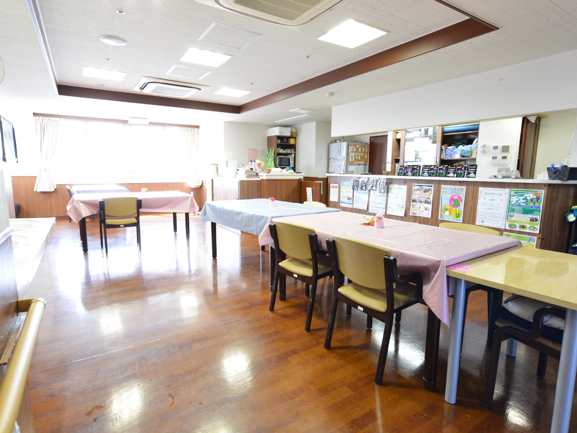 焦げ茶色のフローリングで、白い天井と白と木目調のコンビネーションの壁。中央にはテーブルと椅子が並べて置かれている。横側にはカウンターがある。