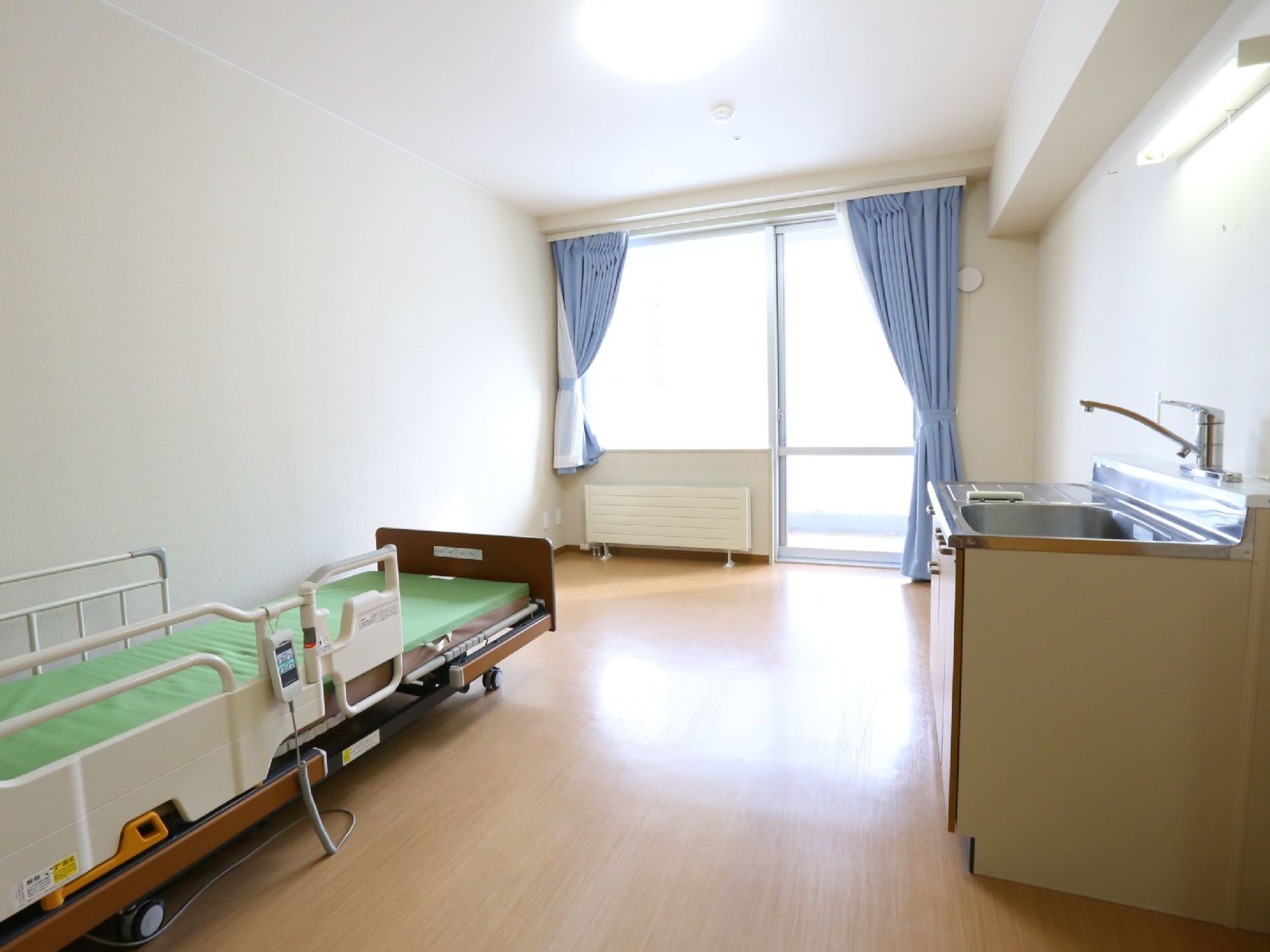 大きな窓があり、陽当たりの良い居室。壁際にはベッドが置かれている。反対側には流し台が設置されている。