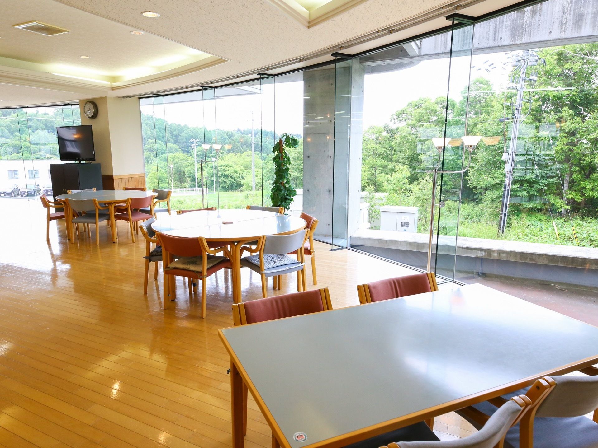 壁一面ガラス張りの明るい食堂である。丸テーブルや長方形のテーブルが置かれている。柱の前にテレビが設置されている。