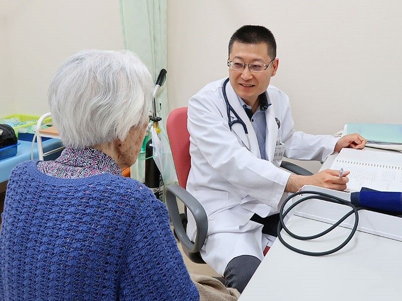 眼鏡をかけた男性医師と高齢女性が花している様子。医師は白衣を着ていて、ボールペンでなにかを書いている。