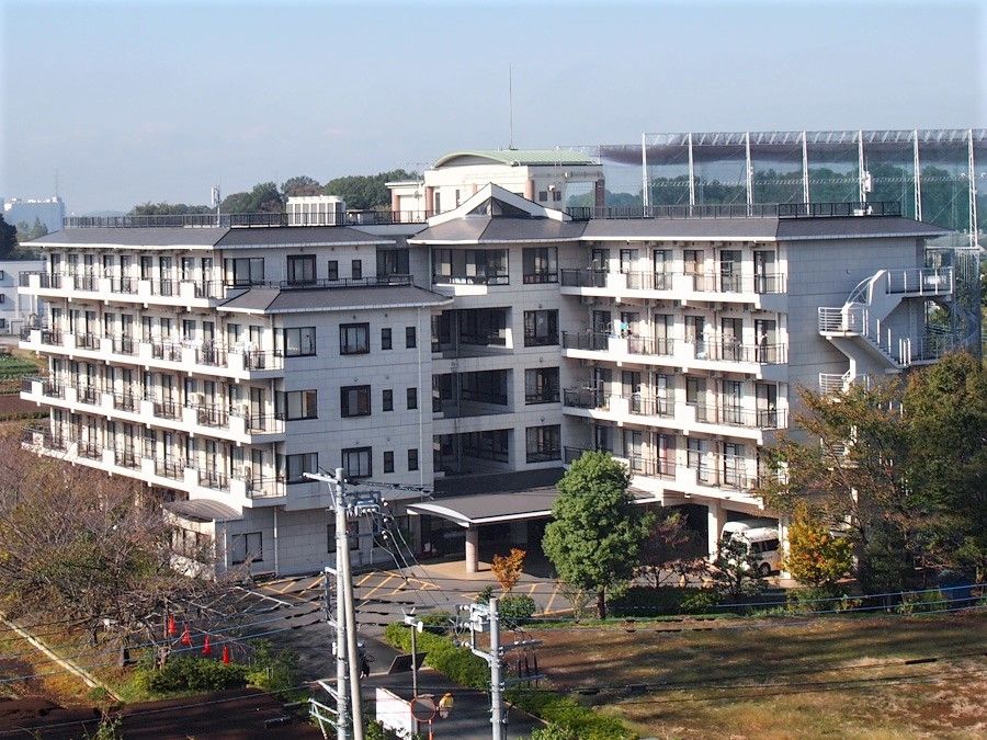 ５階建ての建物は白を基調とし、周辺には緑が多く、駐車場もある。