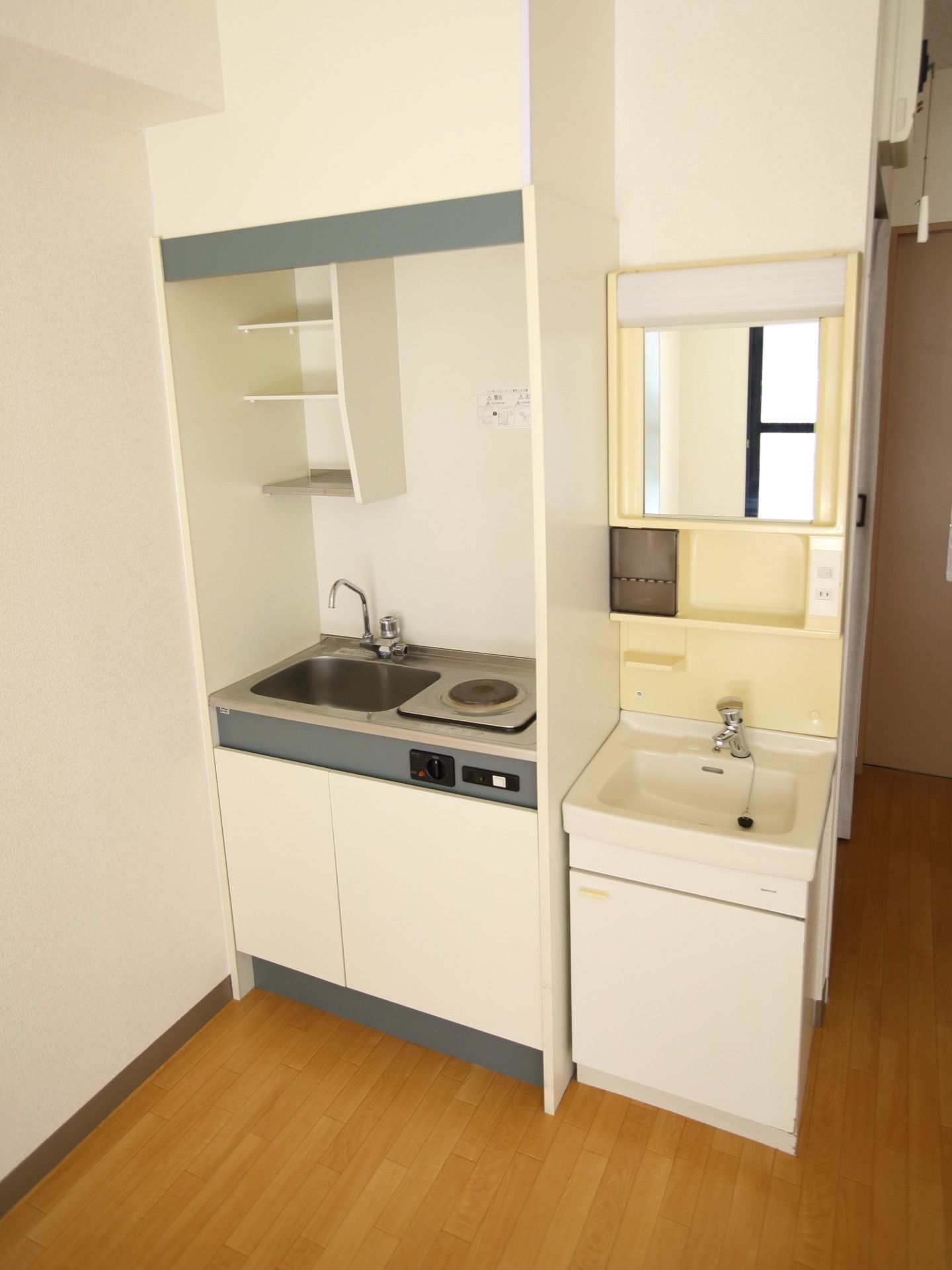 居室にはミニキッチンと洗面台が並んで設置されている。キッチンには電気コンロが付いており、流しの上には棚が設けられている。