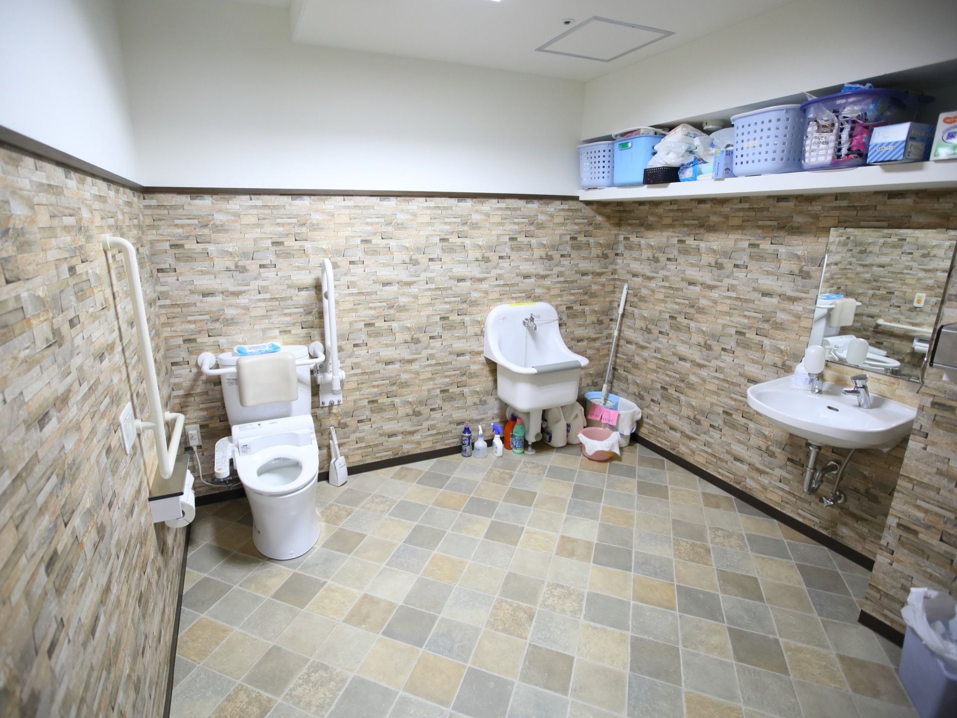 トイレはとても広い空間で床にはさまざまな色のタイルが張られている。左側には温水洗浄便座があり、右側には洗面台がある。