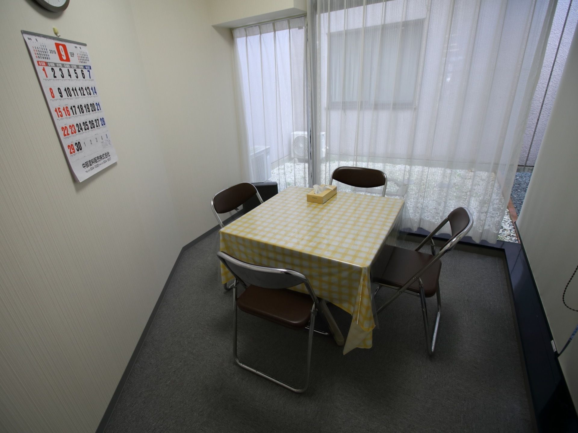 グレーのカーペットが敷き詰められた、落ち着いた雰囲気の談話室。中央にはテーブルと椅子が置かれており、壁にはカレンダーが掛けられている。