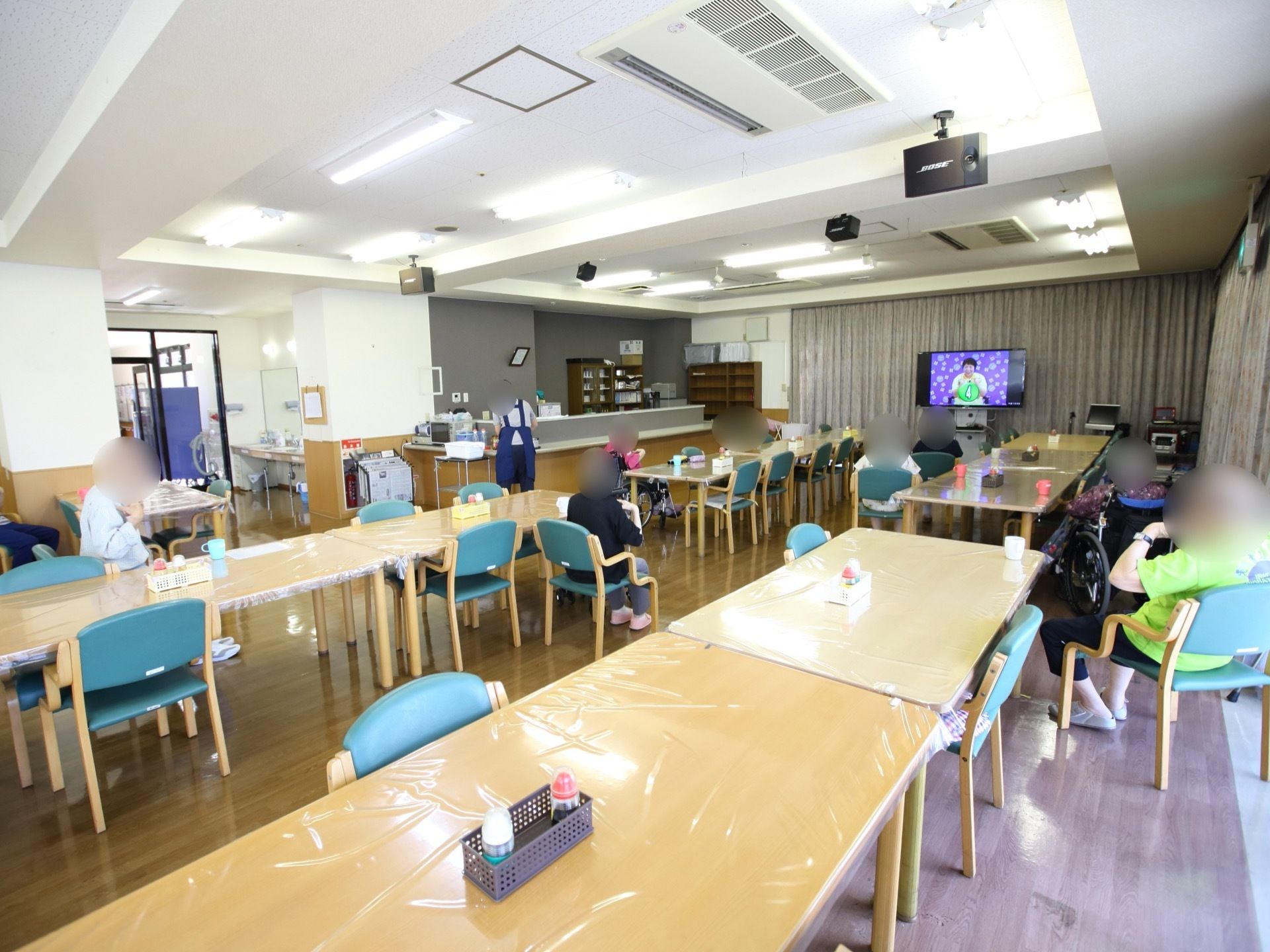 食堂にはテーブルが並べられており、入居者様が奥にあるテレビに向かって椅子に座っている。天井には空調やスピーカーがある。
