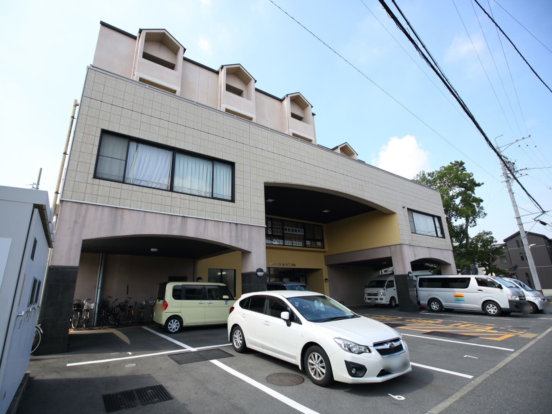 建物中央の奥まった場所に玄関があり、上には大きな屋根が付いている。建物の前には駐車場が設けられ、車が数台停まっている。