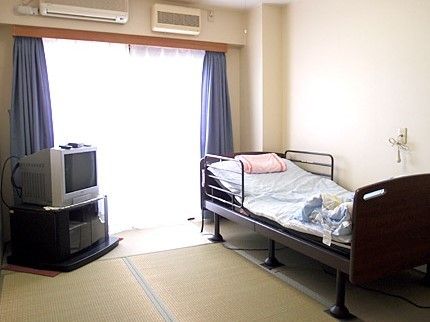 和風の居室。左手のベッドサイドにはナースコールがあり、右手にはテレビ台＆テレビ、窓からは明るい日差しが降り注ぐ。