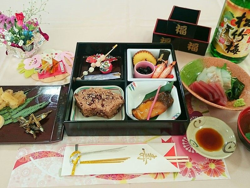 天ぷらや刺身、赤飯、ぶりの照り焼きなどが並んだ食事。そばには寿と書かれた割りばしや日本酒もある。