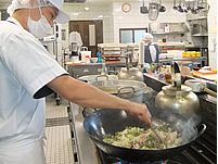 白いマスクや帽子、調理用の服を着たスタッフが大きな中華鍋を使用して料理をしている様子。