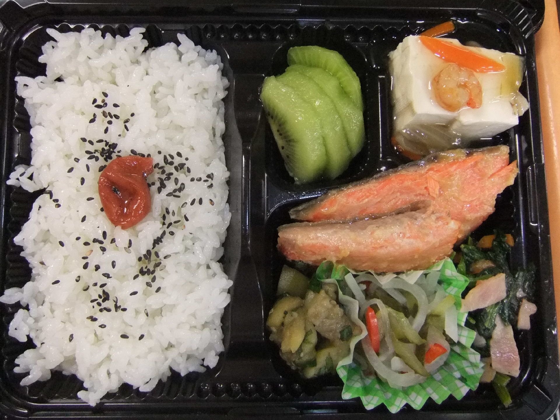 黒い入れ物に入ったお弁当。白ご飯の上には梅干しと黒ゴマ、酒や野菜のあえ物、豆腐のあんかけ、キウイなどが盛られている。