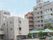 屋外階段が付いたハウスは大きな建物で、すぐ隣には医療機関がある、共にベージュ系の外壁で、駐車場がある。