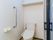 白を基調とした清潔感のあるトイレである。シルバーの手すりがある。仕切りにじゃばらのカーテンを使用している。
