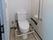 トイレの便座のそばには、手すりが配置されている。白い壁の上部には収納棚が設置され、トイレットペーパーが１つ置かれている。