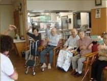 いすに座ったママ体操に励む入居さの様子。前でスタッフがお手本を見せている。