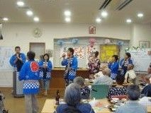 入居者は席につき、はっぴを着たスタッフがマイクを持って歌を歌っている様子を眺めている。
