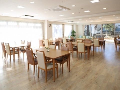 大きな窓が複数ある明るい空間には、入居者様のために４人がけのテーブルが置いてある。椅子には２つのデザインがあり、互い違いに配置している。