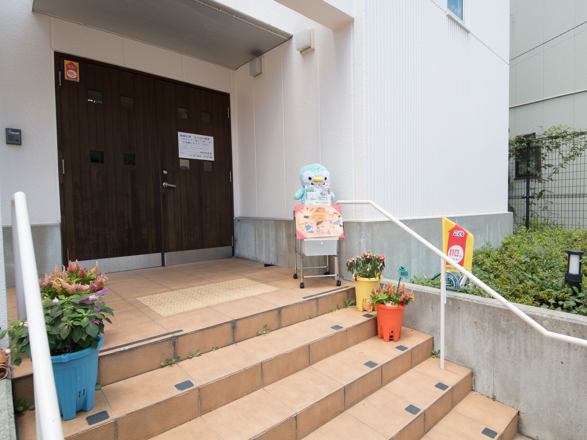 玄関前の階段には手すりがあるので安全に昇り降りができる。階段の手前には転落防止用の点字ブロックがある。季節の花が飾られている。