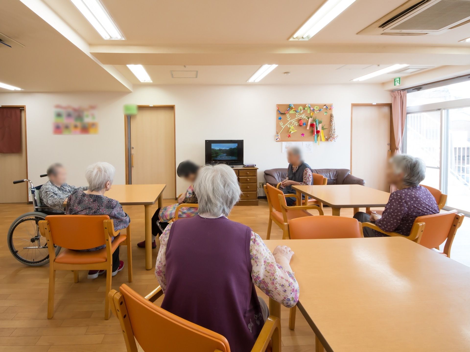 大きな窓のついた明るい談話室。広々とした部屋には４人掛けのテーブルセットが３つとソファが置いてある。壁に季節の飾りが飾られている。