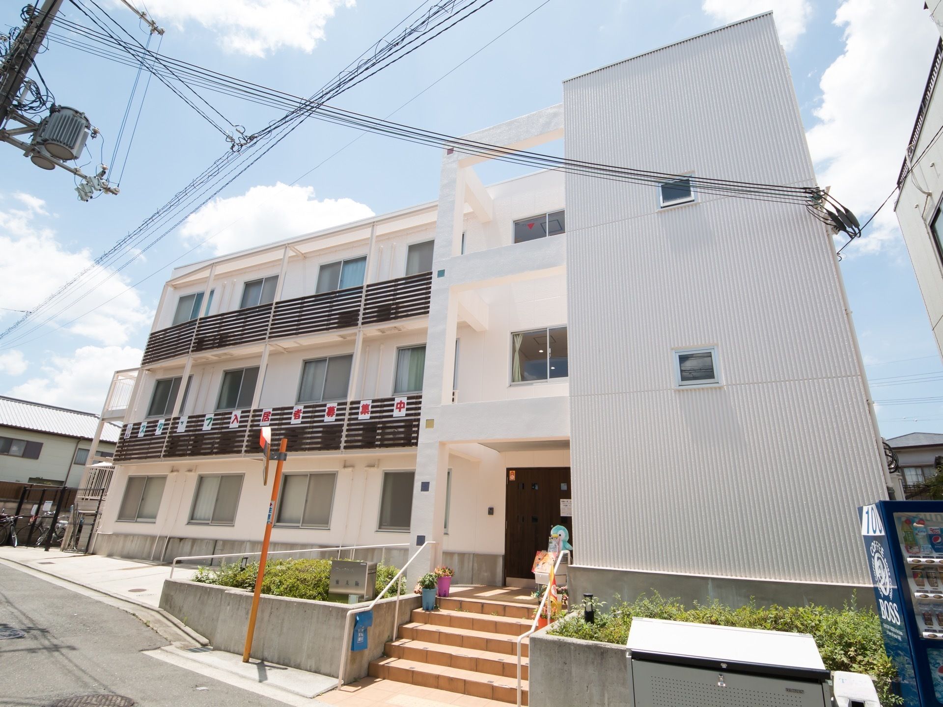建物の前には自動販売機や、金属製のダストボックスが置かれている。左端には自転車が置けるスペースが設けられている。