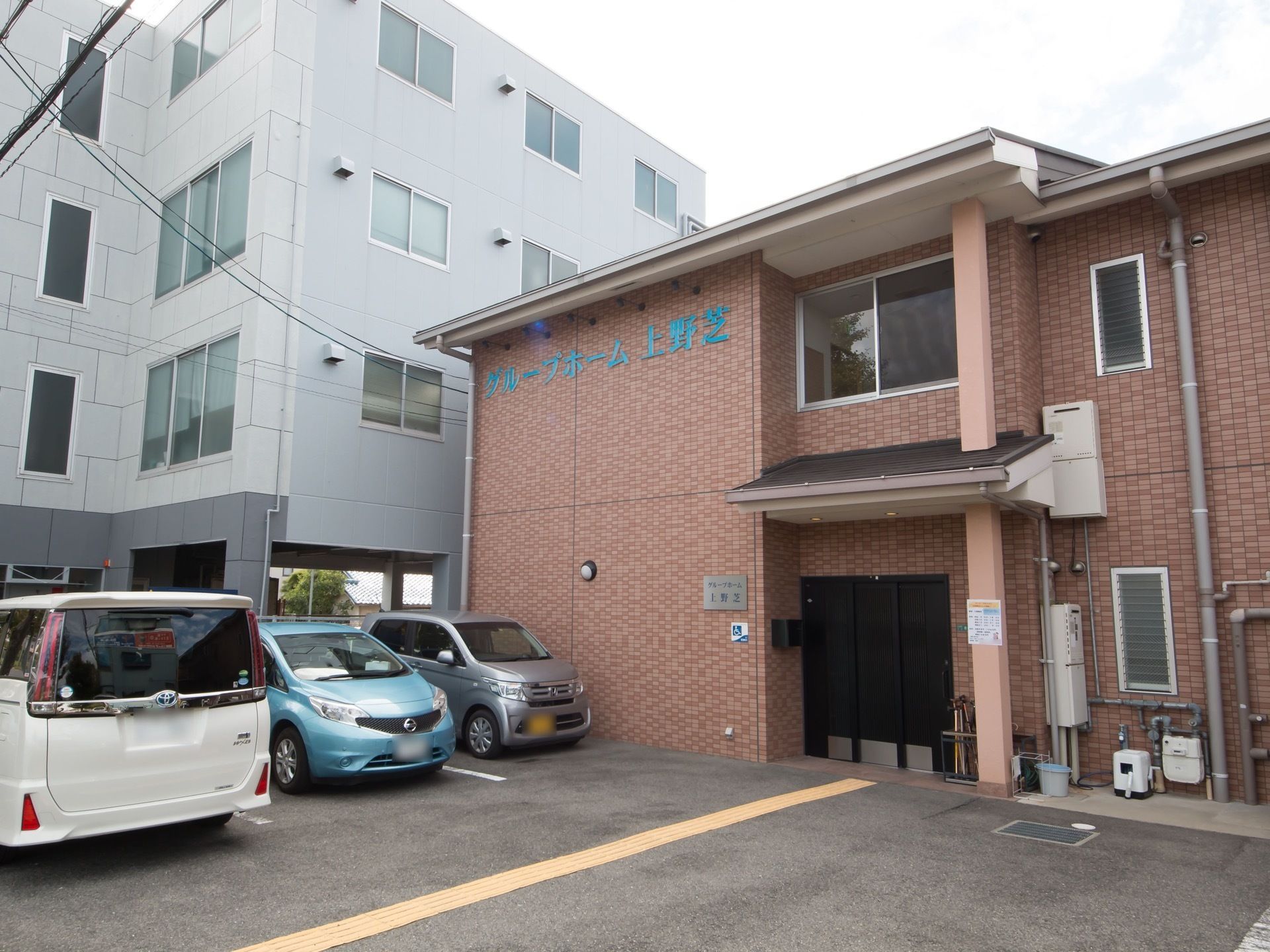 落ち着いた色合いの外観である。２階部分にブルーで施設名が描かれている。建物の前には、駐車場が完備されている。