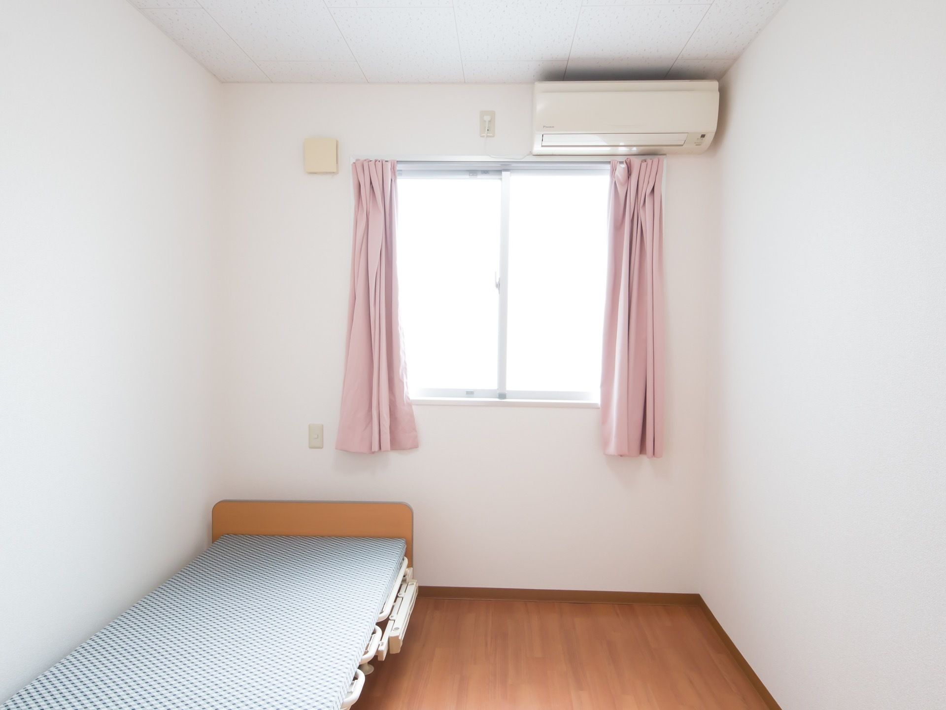 白い壁にエアコンが設置され、大きめの窓にはピンクのカーテンが取りつけられている。ベッドが１台置かれている。