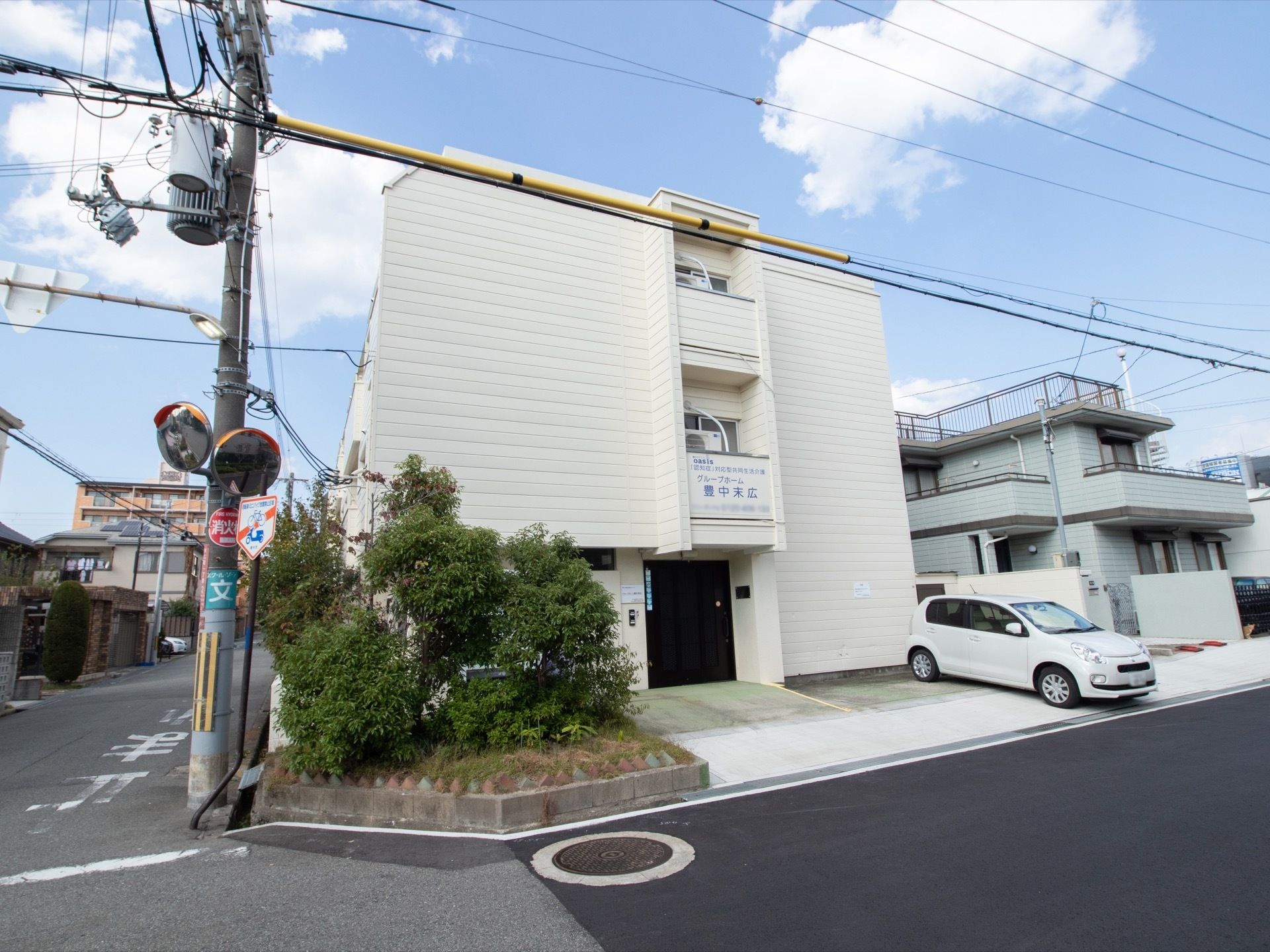 施設は３階建てなっており、住宅街に囲まれている。右側に駐車場が完備されている。扉の真上に看板が設置されている。左側には木が植えられている。