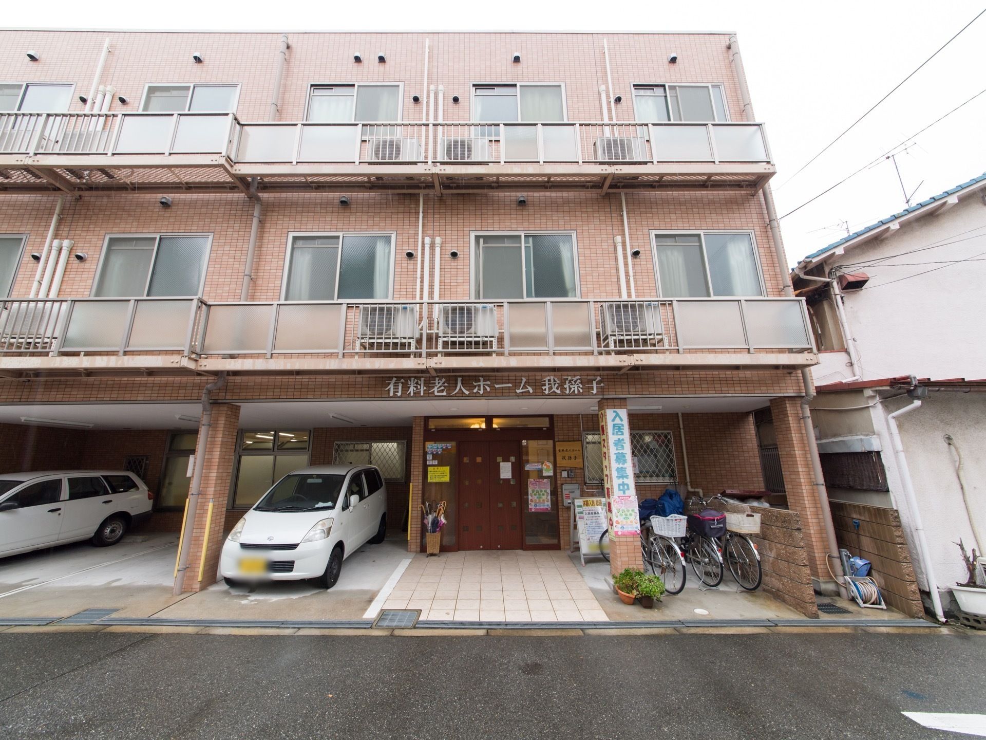 駐車スペースには２台の自動車が停車中である。傘立てには傘が数本立ててある。正面に大きく施設名が表記されている。