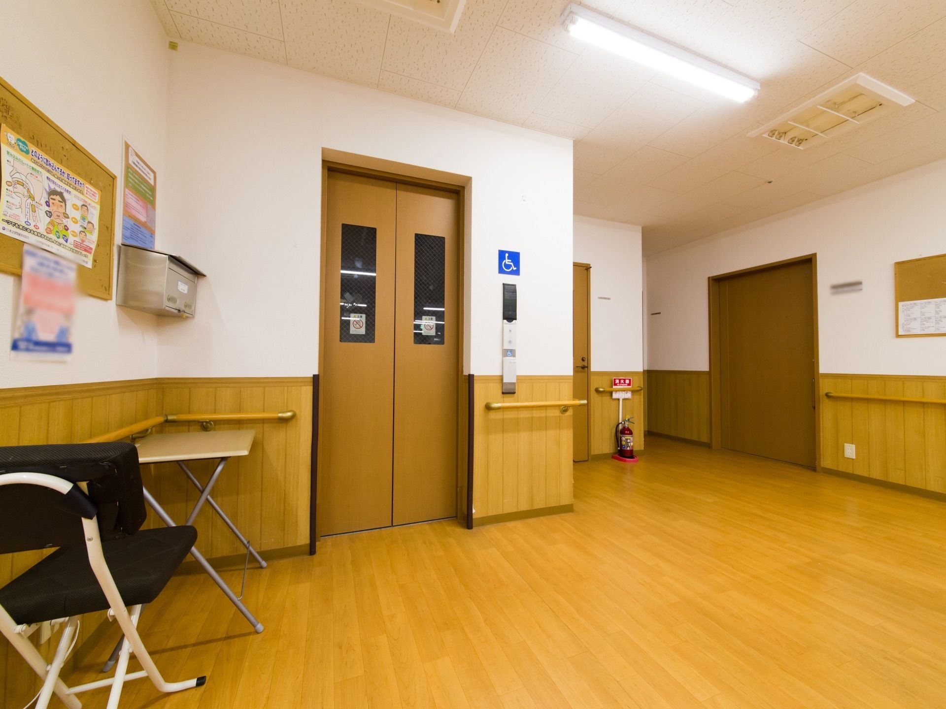 車いすで利用が可能なエレベーターである。出入り口付近には小さなテーブルとイスがある。手すりが設置されている。壁には掲示物が貼付されている。