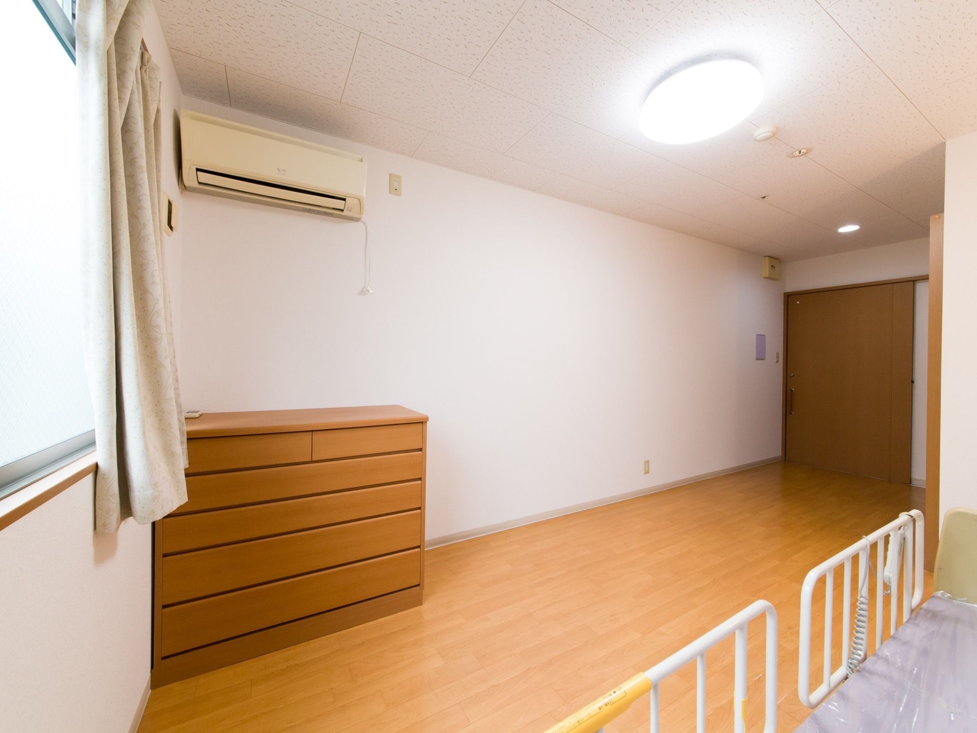 木目調の床と白い壁面の内装である。出入り口は引き戸である。エアコンが設置されている。備え付けのタンスと介護用ベッドがある。