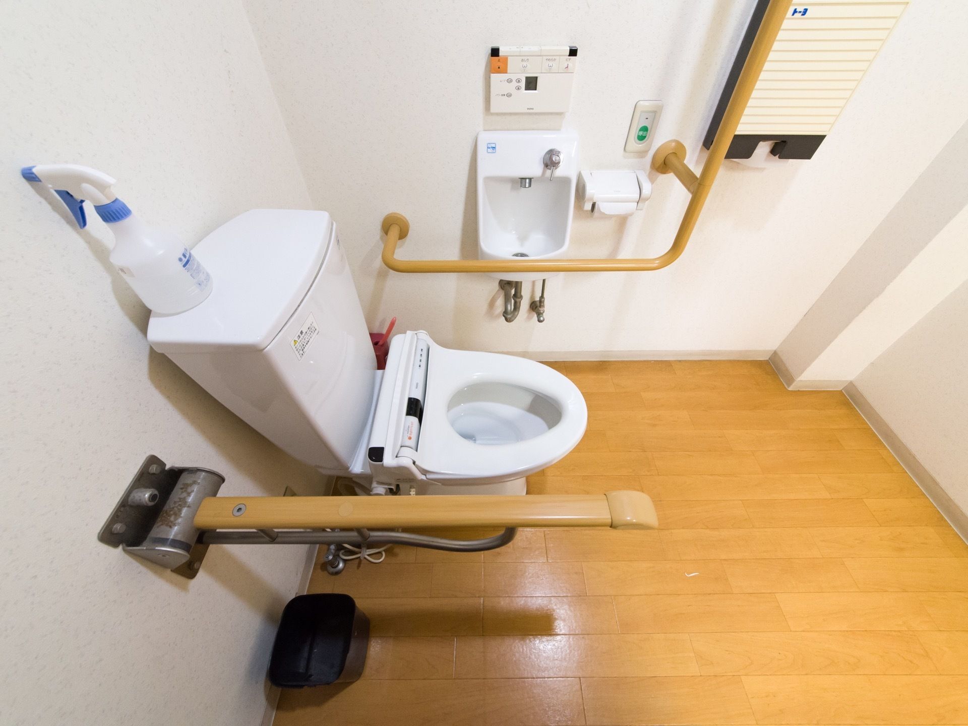 床は木目調である。便器周辺には手すりを設置している。非常用呼び出しボタンがある。ごみ箱の用意がある。