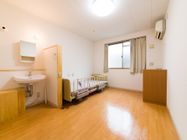 木目調のフローリングの居室である。窓にはカーテンが取り付けられている。エアコンと介護用ベッドが設置されている。