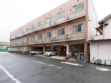 茶色い外観である。自動車が２台停まっている。３階建ての建物である。出入り口に「入居者様募集中」という表記がある。