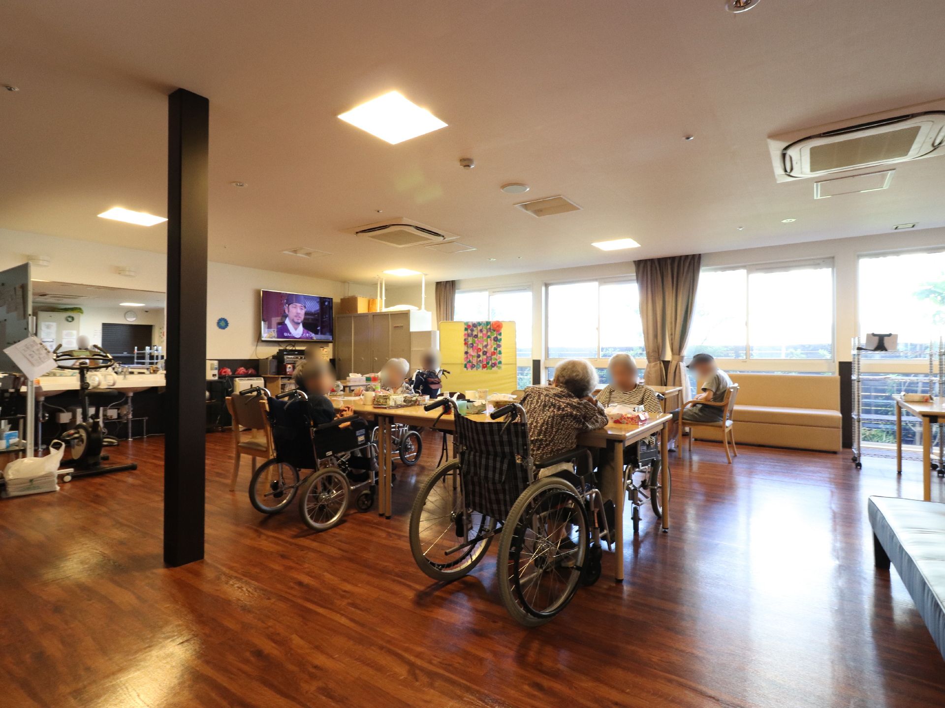 床は木目調のダーク色に黒色柱が立っている。入居者が食事している。その車椅子は広めに間隔を空けられている。