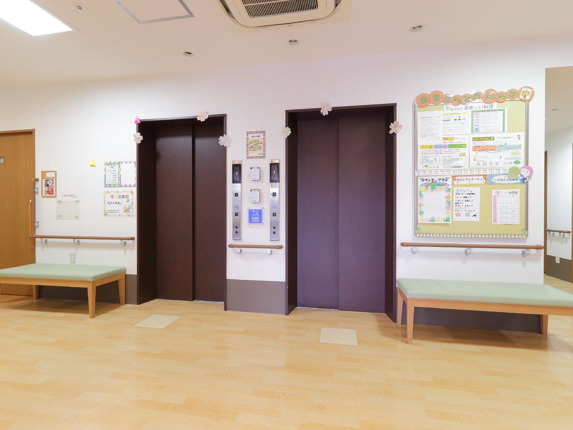 焦げ茶色のシックな扉のエレベータの上部には、花飾りが付いている。壁には手すりが、また付近にはベンチがある。