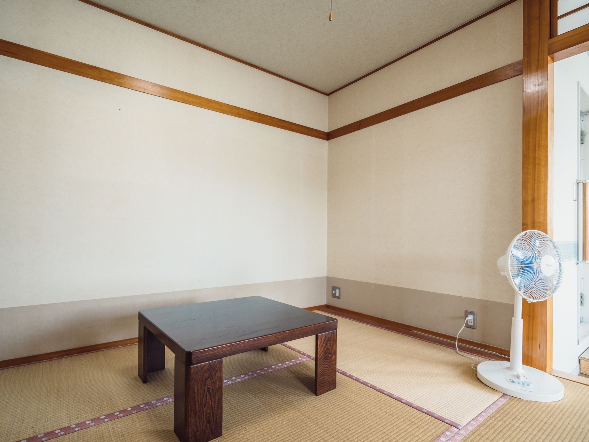 居室は畳敷きの和室である。四角い天板の木のテーブルが部屋の中央に置かれている。壁際には、電源コードがつながった扇風機がある。
