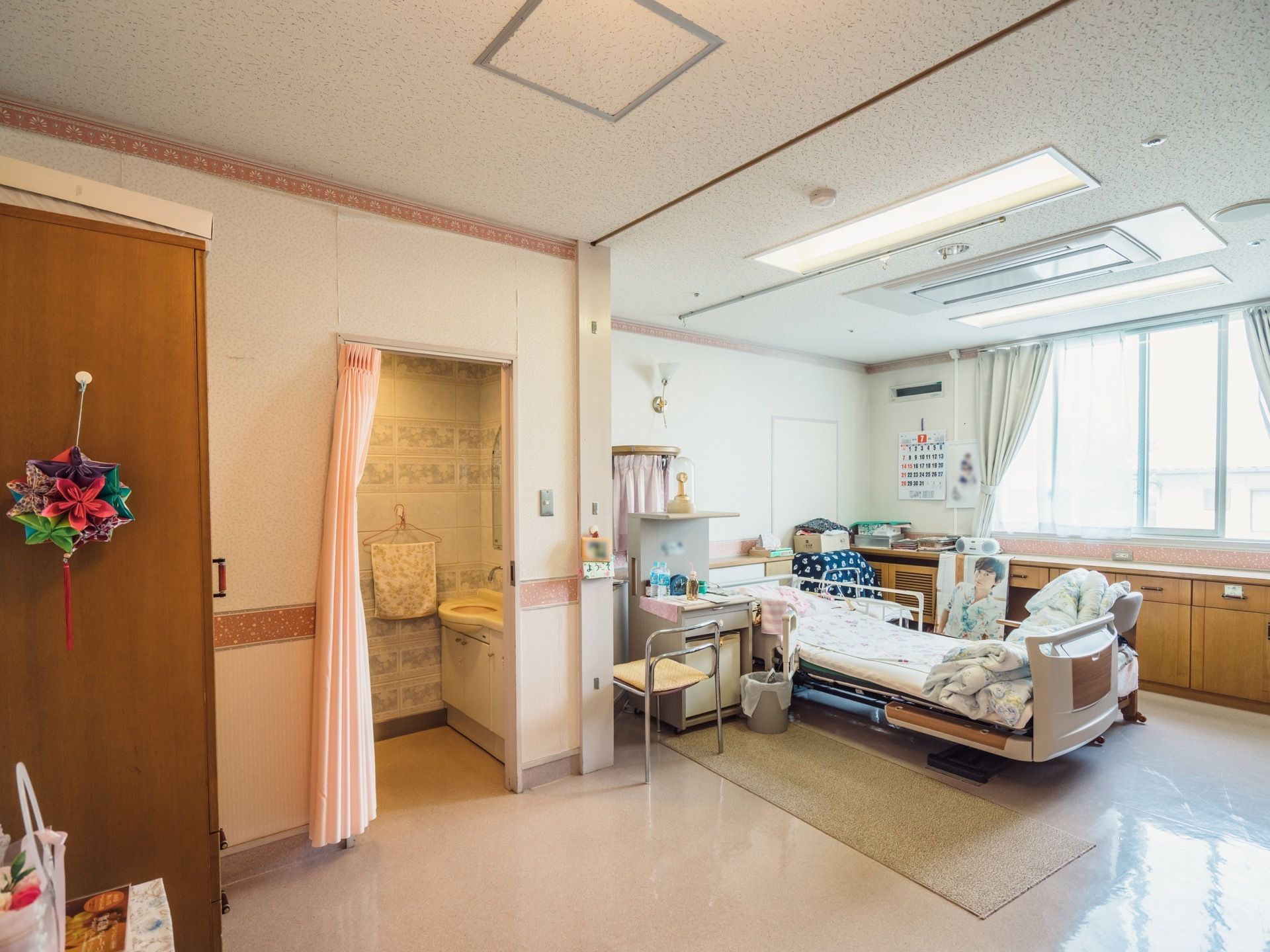 居室には大きな窓がある。転落防止柵がついた介護ベッドの足元に横長のマットが敷かれている。男性アイドルのポスターがある。