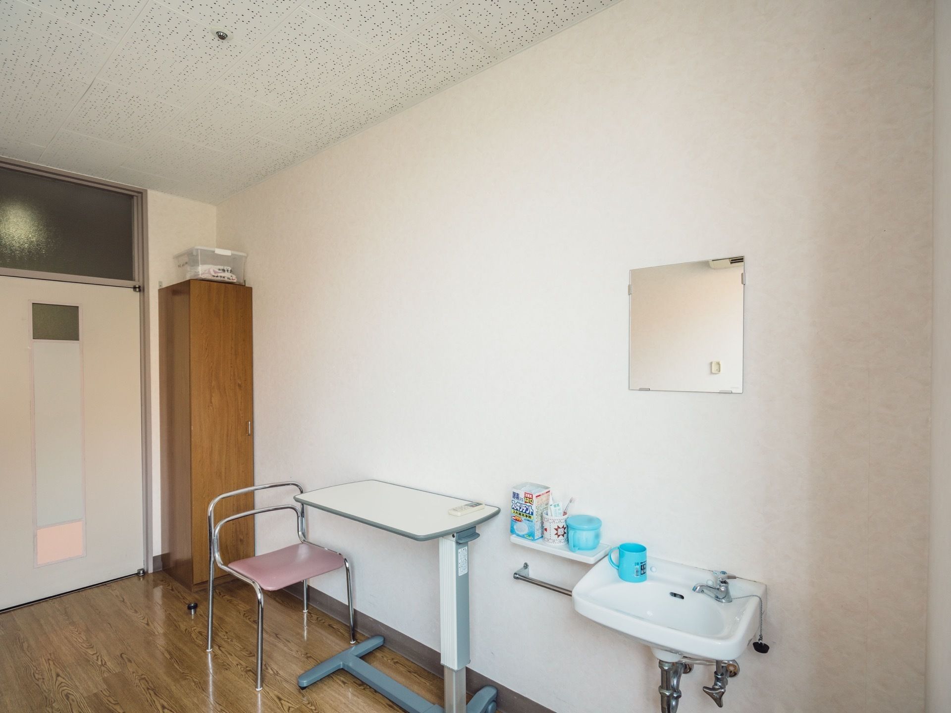 居室に、可動式のテーブルと椅子が置かれている。壁設置式の洗面台があり、コップが載っている。その横には小さな棚がある。