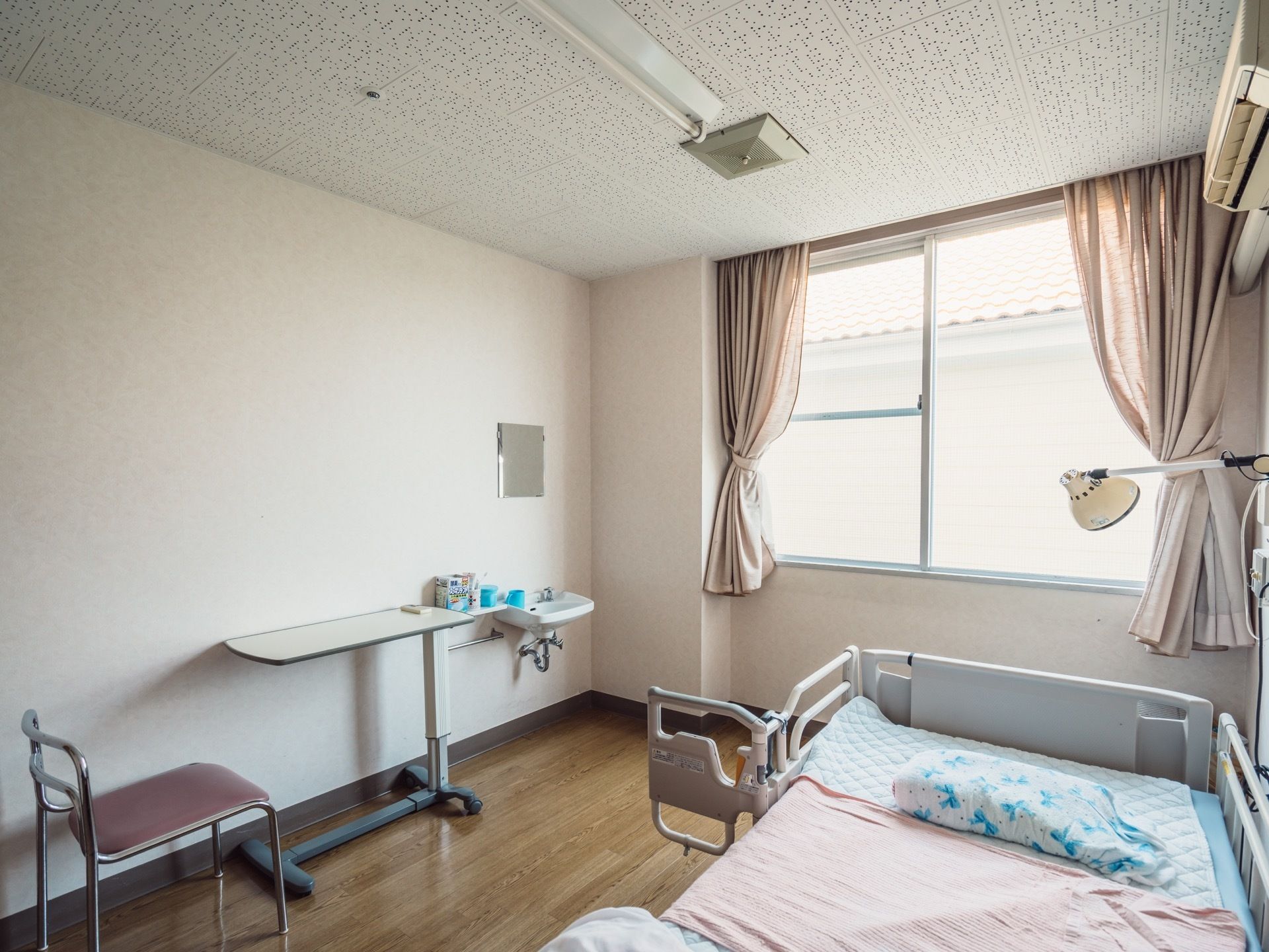 居室には大きな窓があり、ピンク系統の色のカーテンが束ねられている。転落防止柵つきのベッドが置かれていて、その上にエアコンが設置されている。