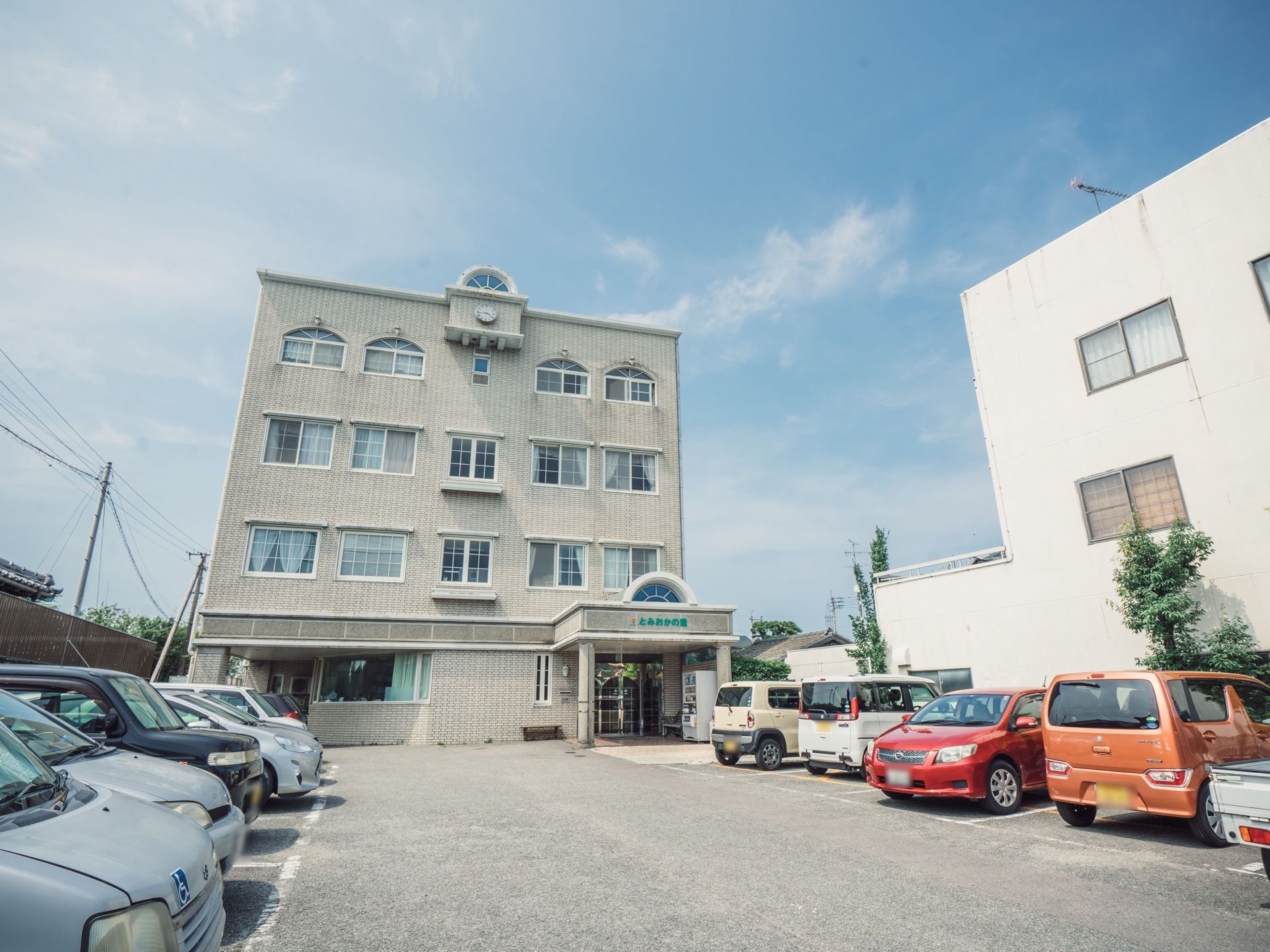 白色を基調とした外壁の４階建ての施設である。敷地内に白色の線で囲まれた駐車スペースがあり、多くの車が停まっている。