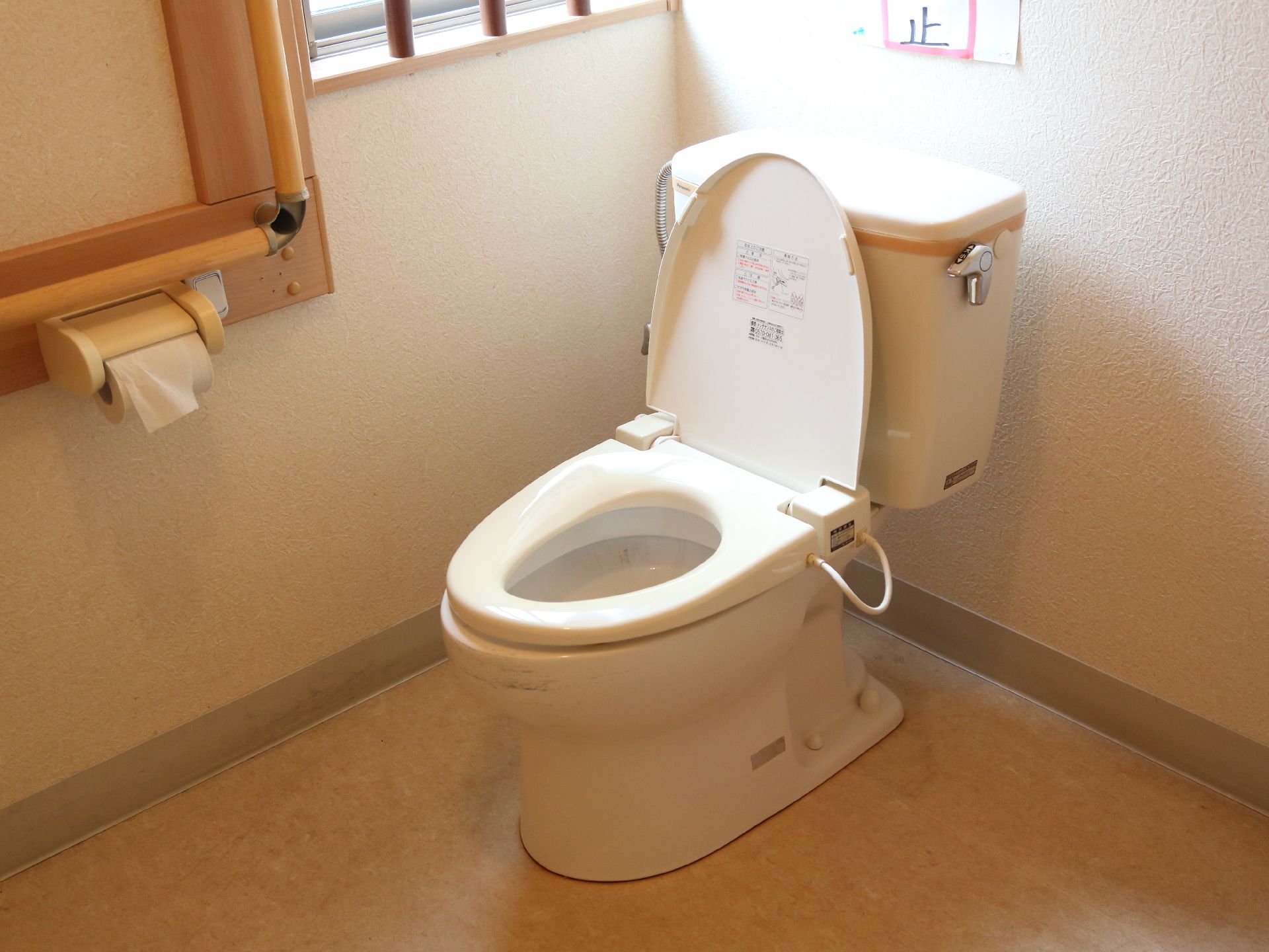 タンク式の水洗洋式便器がある。タンクに水を流すレバーが付いている。右側にトイレットペーパーがあり、L字に手すりが付いている。