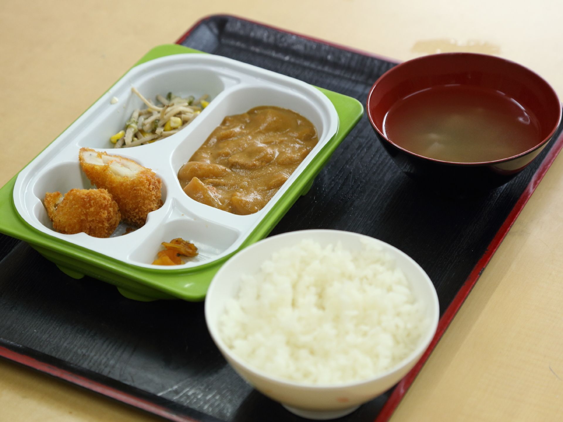 黒いお盆の上にご飯とみそ汁とワンプレートに盛り付けられたおかずがのっている。カレーとフライ、もやしの和え物と漬物が入っている。