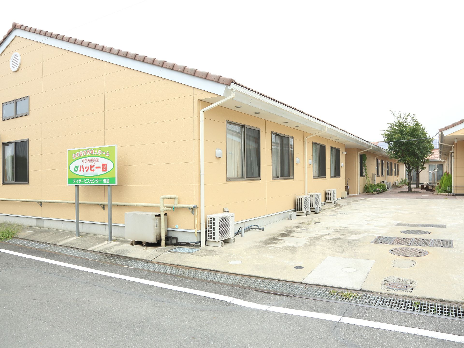 施設は1階建てで瓦の三角屋根になっている。手前に施設名を書いた看板が立ててある。窓の下に室外機が並んでいる。