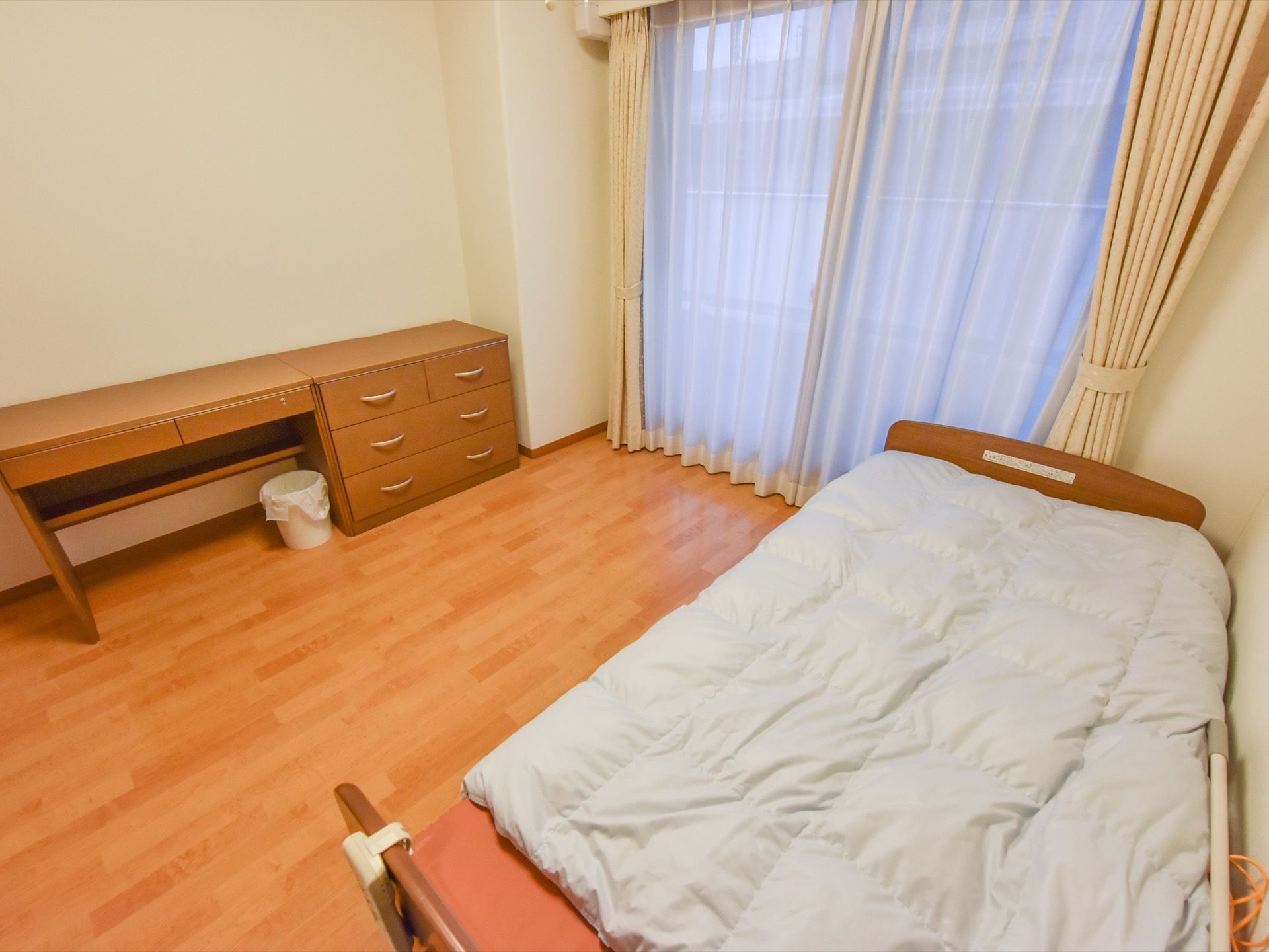 茶色の机と小さめの収納家具が、壁際に並んで置かれている。両方が、同じ高さ、同じ色になっている。白いゴミ箱もある。